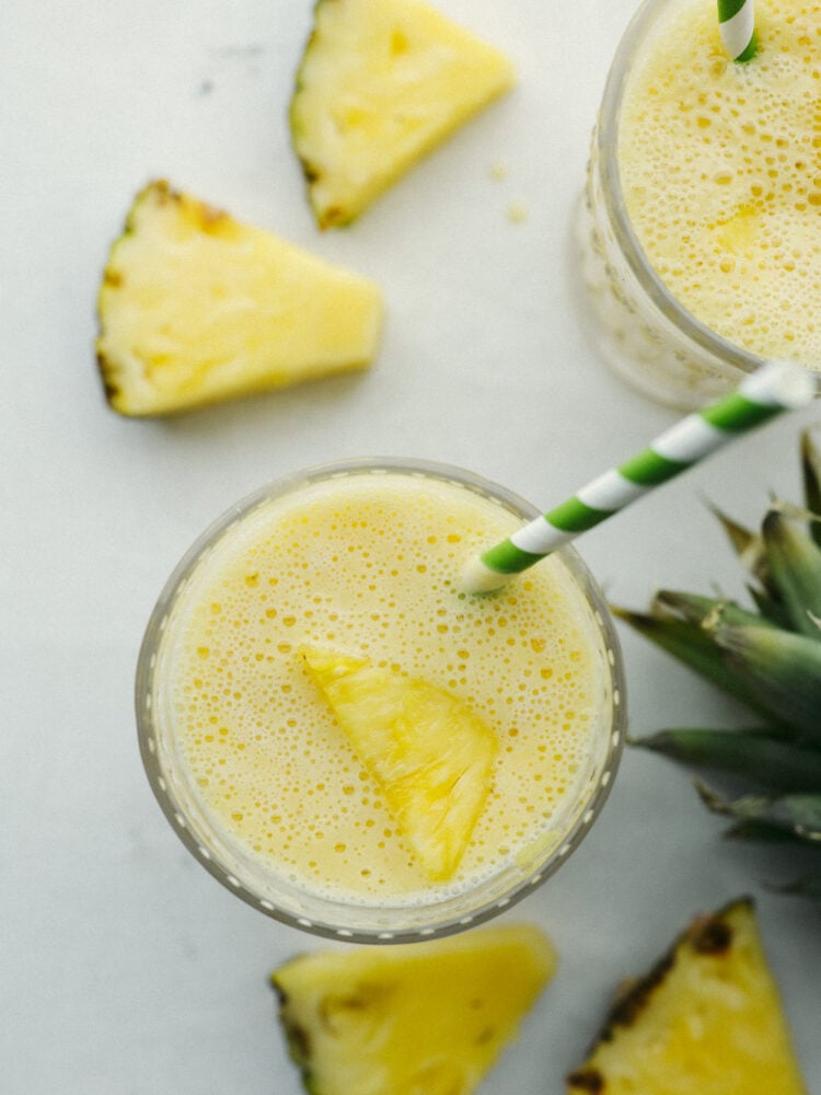 Top view of a pineapple smoothie in a slass with a chunk of pineapple o top and a green and white straw. 