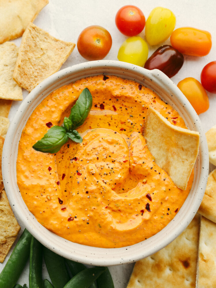 Top-down view of red pepper dip in white bowl, surrounded by chips and peppers.