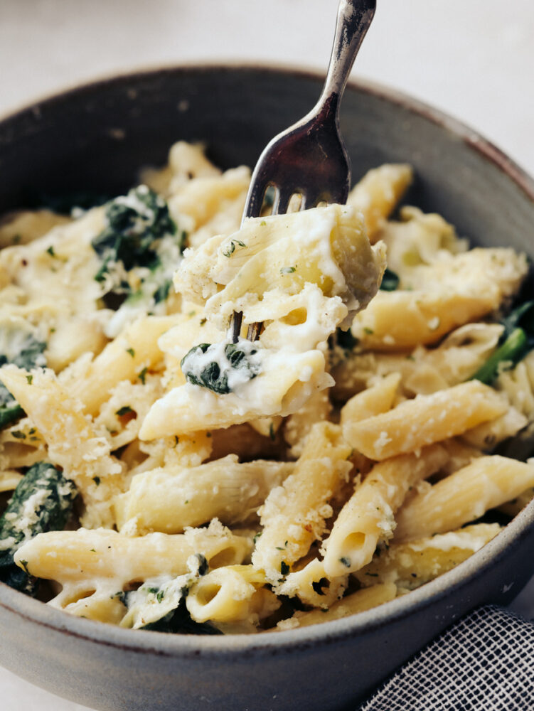 A close up of a bowl filled with spinach artichoke pasta with a fork in it. 