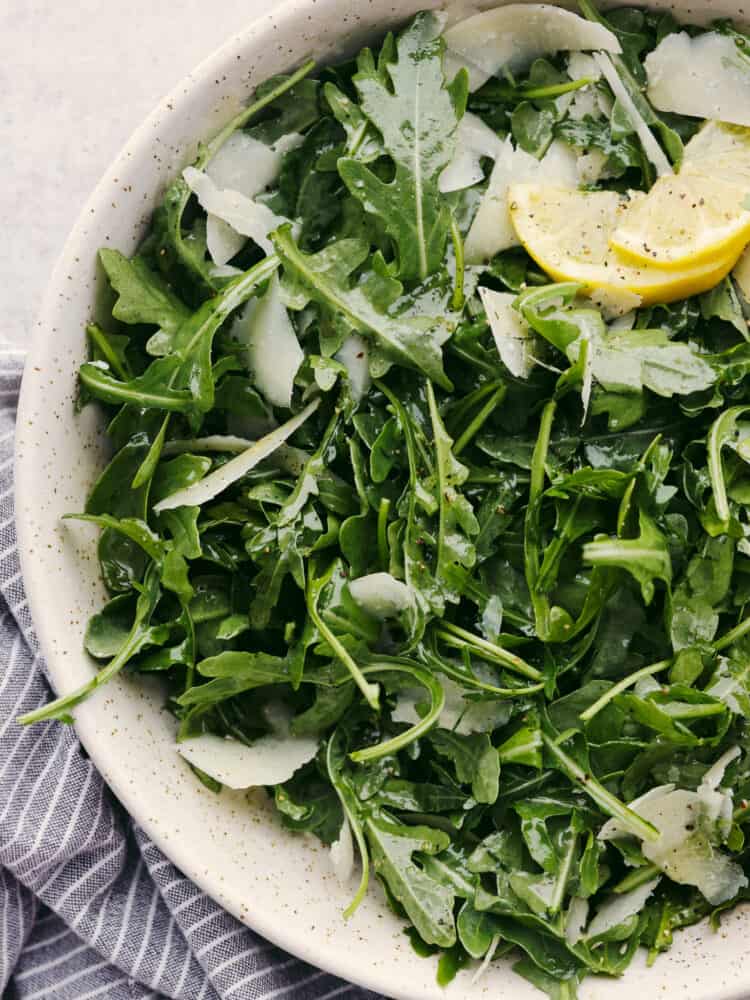 Arugula salad, garnished with lemon, in a white stoneware bowl.