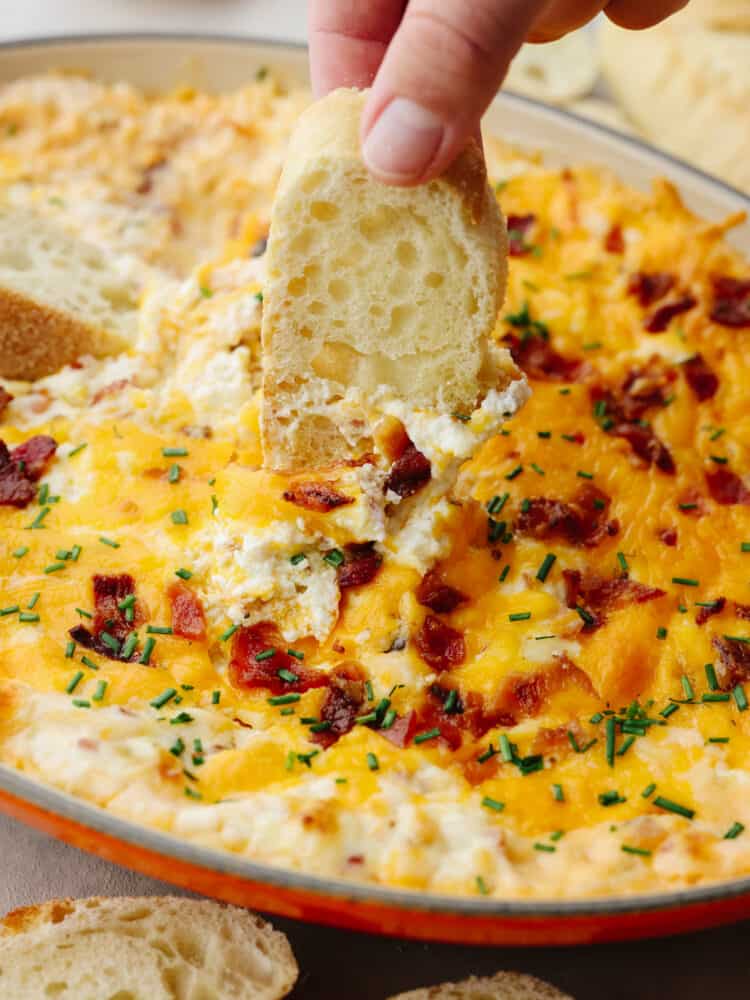 A clice of crostini being dipped into baked bacon dip. 