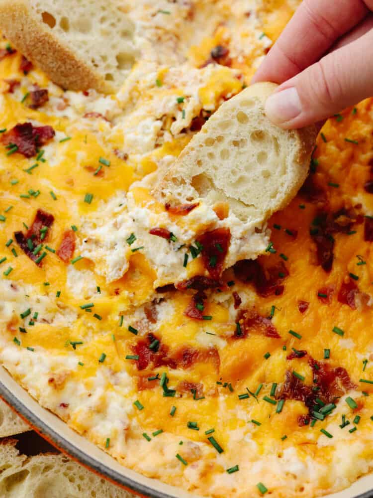 A close up of a slice of french bread being dipped into baked bacon dip. 