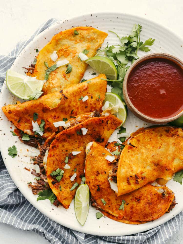 A platter with cooked birria tacos and a small bowl of sauce to dip them in. The plate it garnished with cilantro and lime slices. 