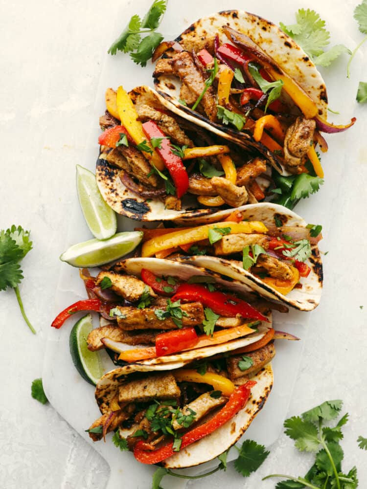 Flour tortillas on a cutting board filled with seasoned sliced pork loin, bell peppers, and onions.  Garnished with limes and cilantro.