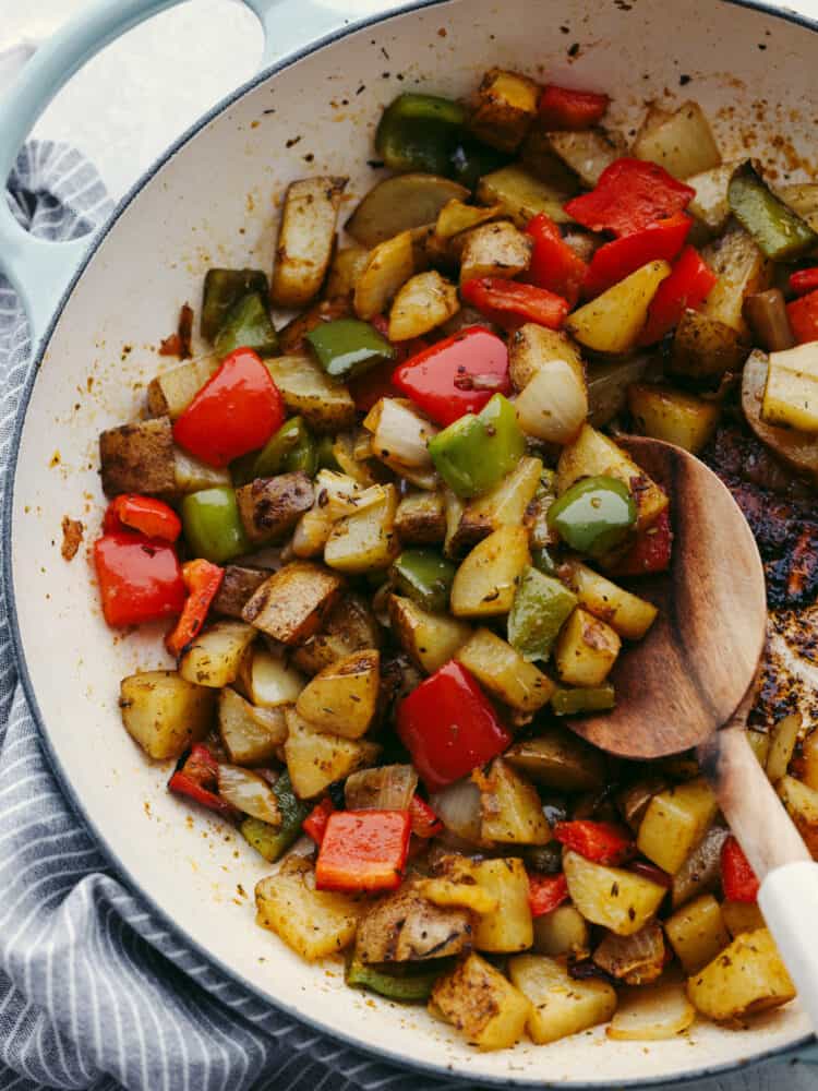 A skillet with cooked potaotes, peppers and onions with a wooden spoon scooping some up. 