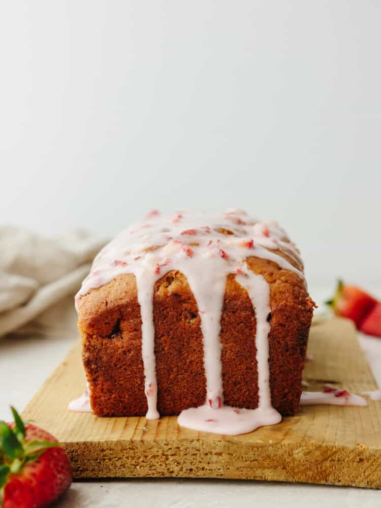 A loaf of strawberry bread on a wooden cutting board with strawberry glae dripping off the top. 