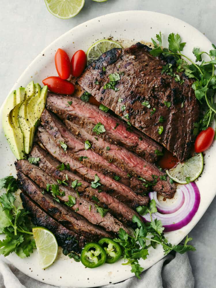 Top view of sliced carne asada on a platter.  Garnished with limes, cilantro, sliced avocado, tomatoes, red onion, and sliced japepeno.