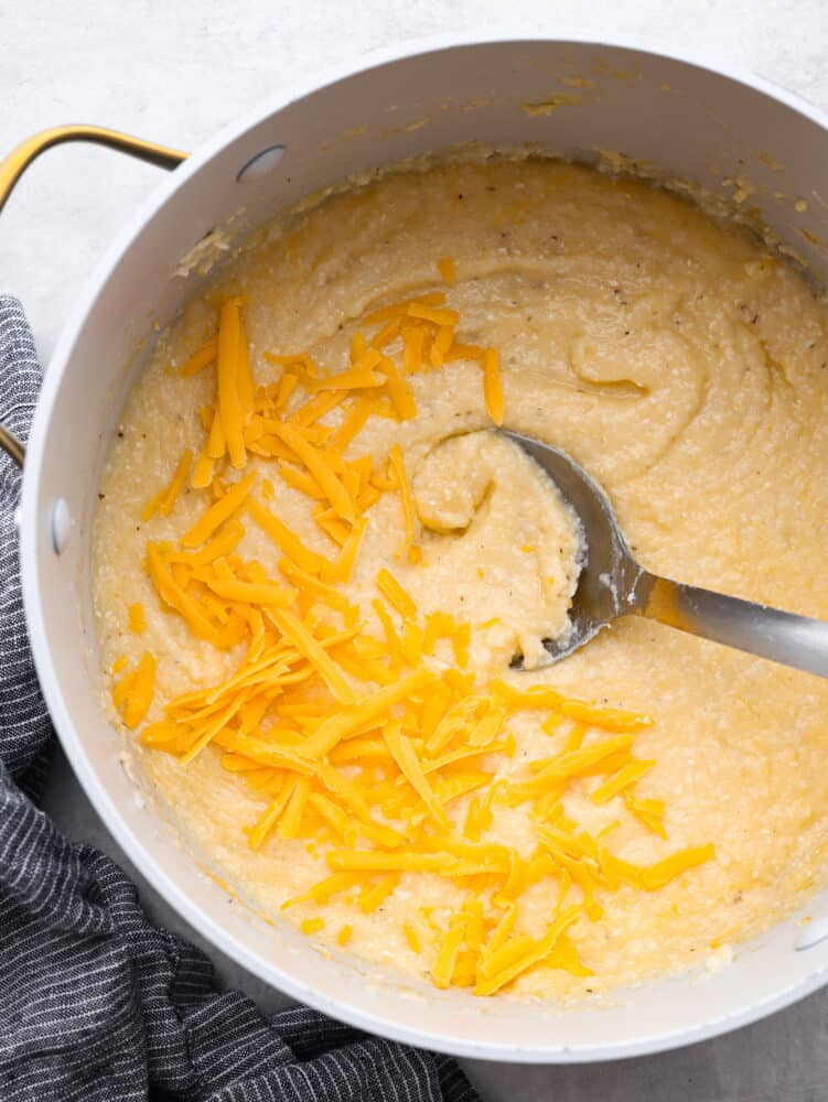 Top view of a large pot of cheesy grits with a large metal spoon laying in the grits.  Shredded cheddar cheese garnished on top with a gray kitchen bowl on the side of the pot.