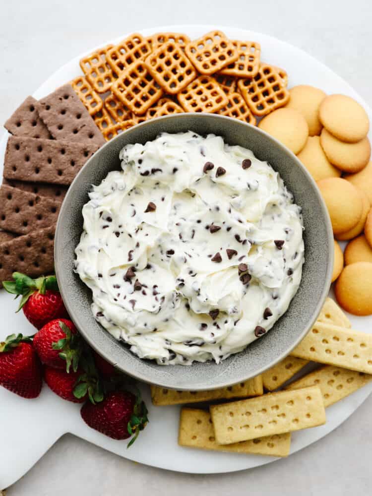 A platter with a bowl of chocolate chip cookie dough dip in a bowl in the middle surrounded by strawberrries, cookie and pretzels. 