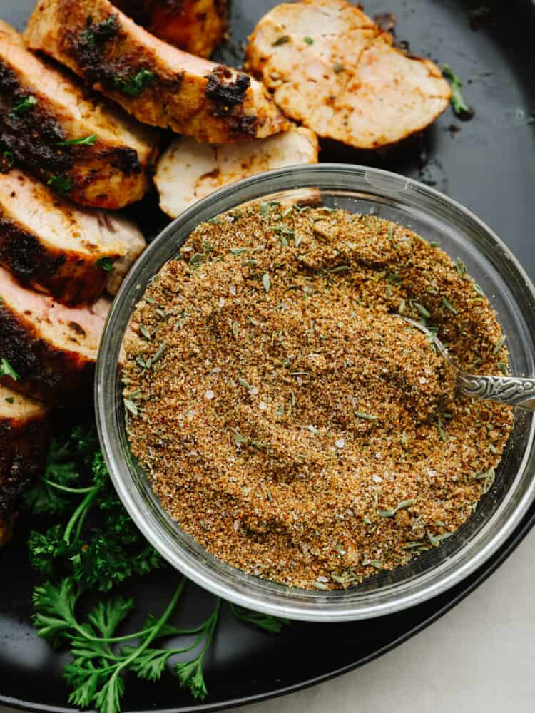 Pork seasoning in a bowl with a spoon on a platter with sliced, cooked pork. 