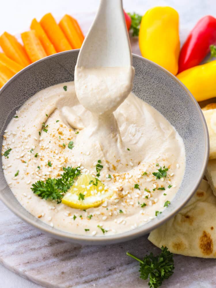 A bowl of tahini sauce on a platter with veggies. A plastic seerving spoon is scooping out some sauce. 