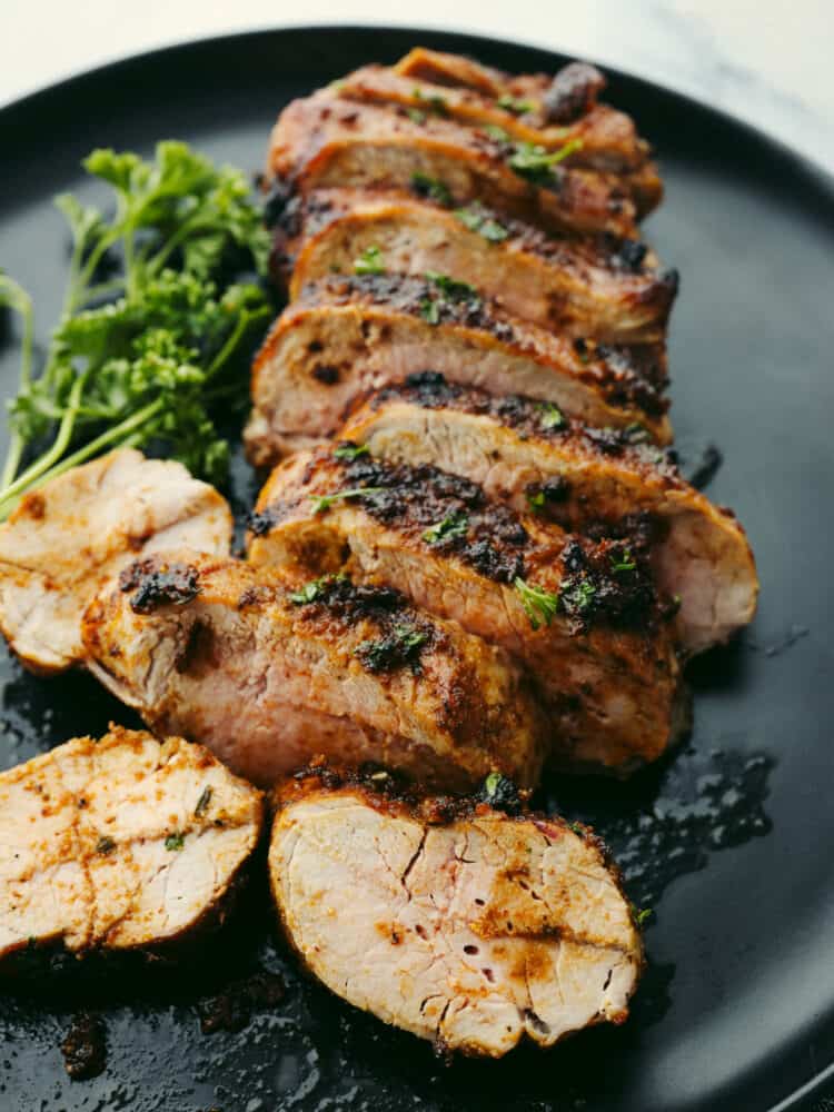 Close up photo of cooked sliced tenderloin on a black plate.  Garnished with a sprig of parsley on the side.