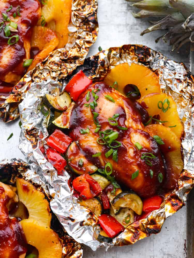 Cooked BBQ chicken in aluminum foil surrounded by cooked pineapple and veggies. 