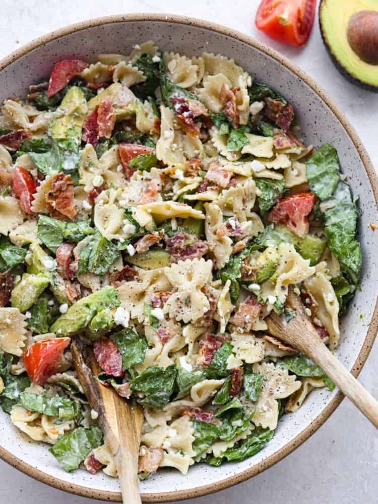 A bowl filled with pasta salad, topped with feta and being tossed with two wooden spoons. 