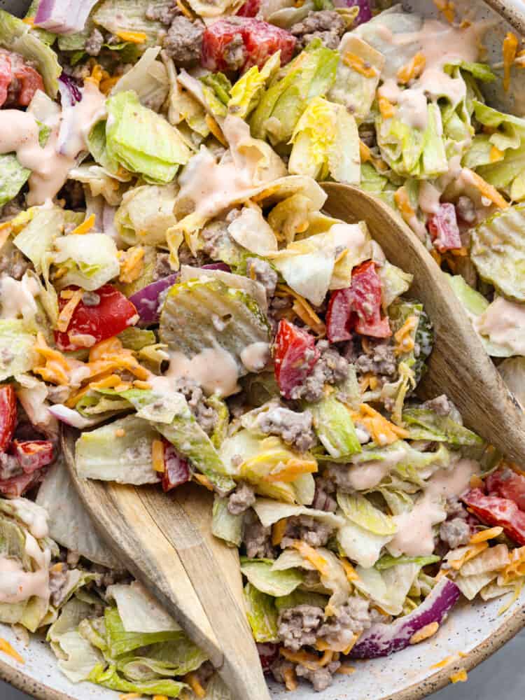 A top view of the tossed cheeseburger salad in a large bowl.  The ingredients and salad dressing are mixed together with wood spoons resting in the salad.