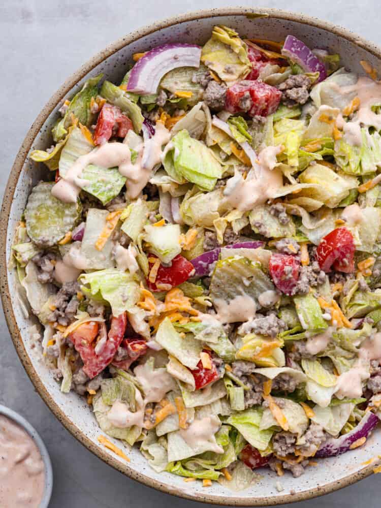 Top view of the cheeseburger salad in a large tan bowl.  The salad is tossed with a thousand island dressing with a small bowl of the dressing on the side.