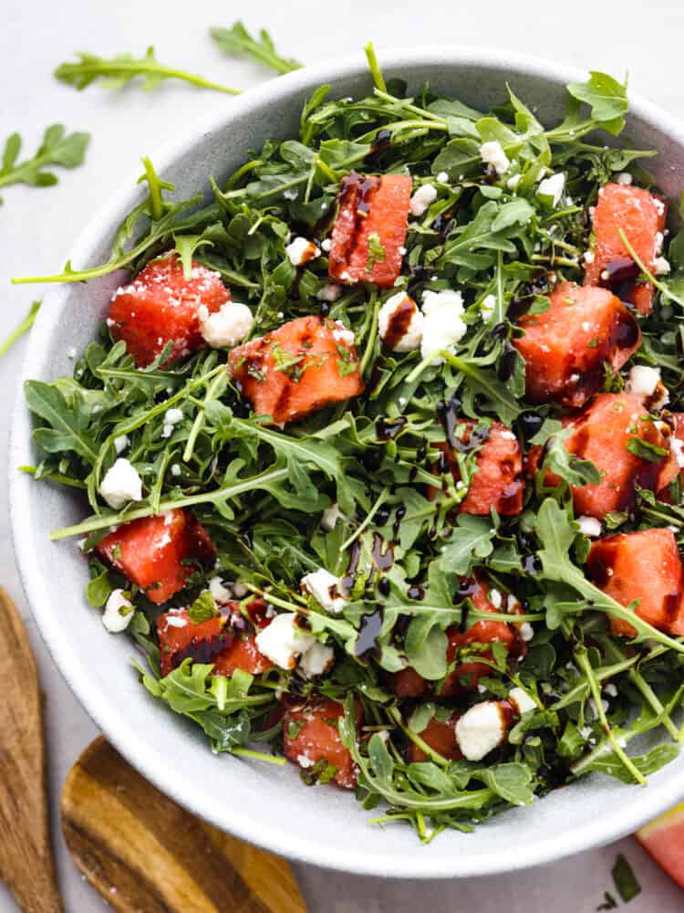 Top view of watermelon arugula salad in a gray bowl.  Wood serving spoons and scattered arugula is laying next to the bowl.