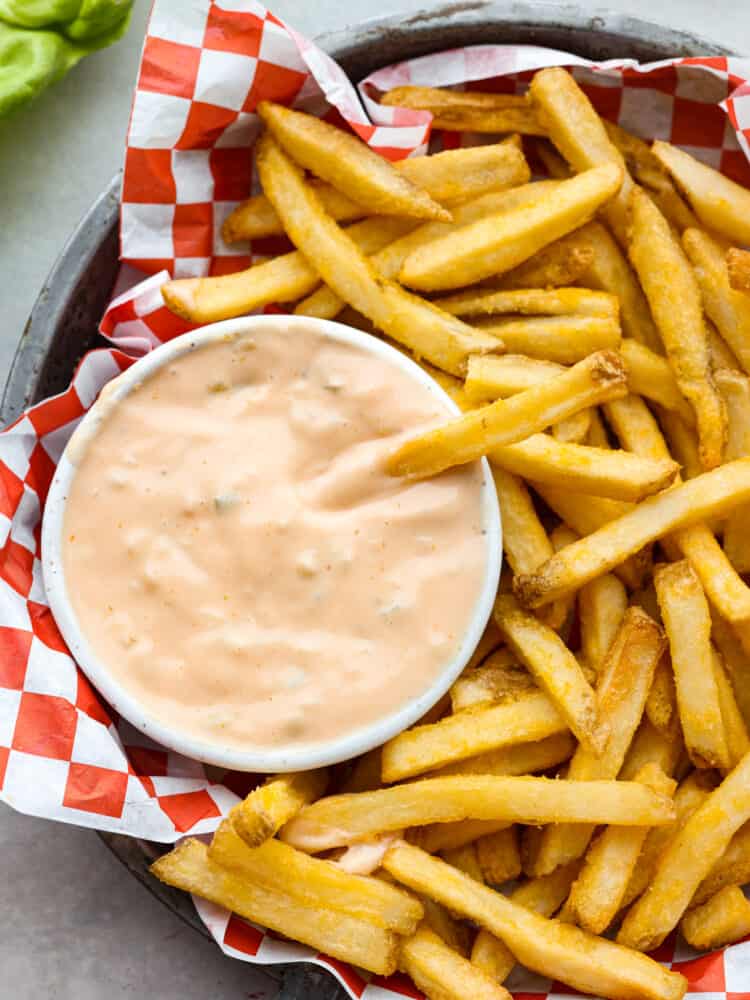 Overhead photo of a French fry dipping in a bowl of sauce. The small bowl of sauce and extra French fries are inside of a metal tin pan.  The metal tin pan is lined with red and white checkered parchment paper.