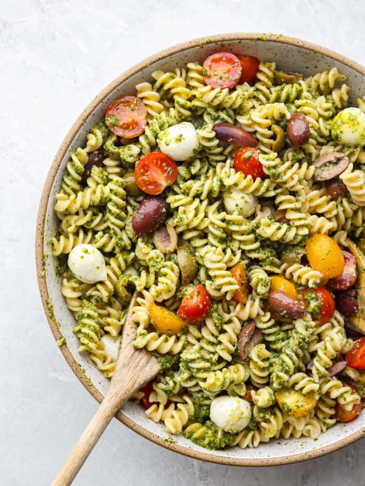 The top view of a bowl with pesto pasta salade and a wooden spoon in it. 