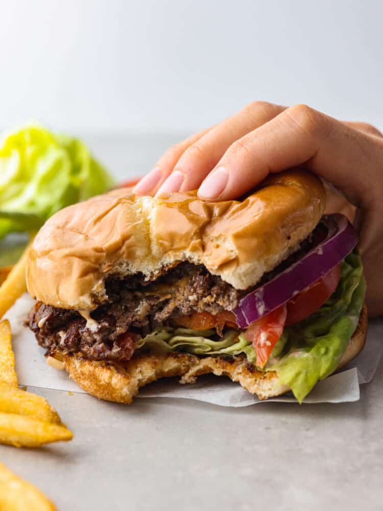 Side view of a hand holding the smash burger with a bite taken out.  Fries scattered on the side.