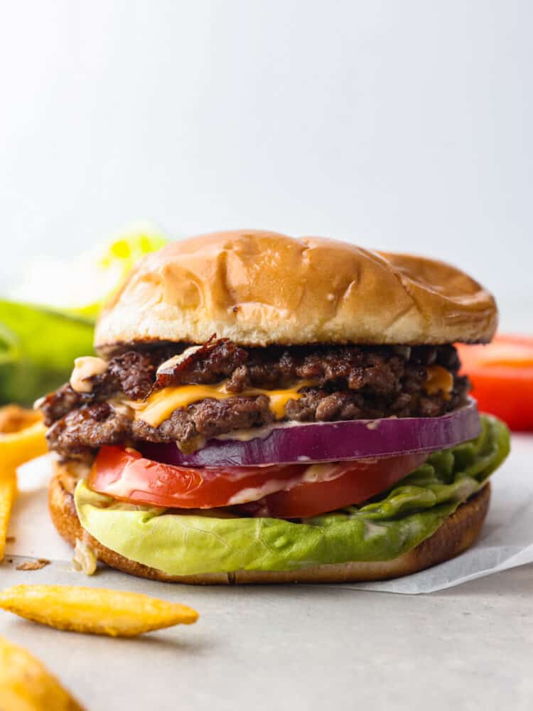 Close up side view of smash burger in a soft toasted bun, and ayered with meat, cheese, red onion, tomato and lettuce.