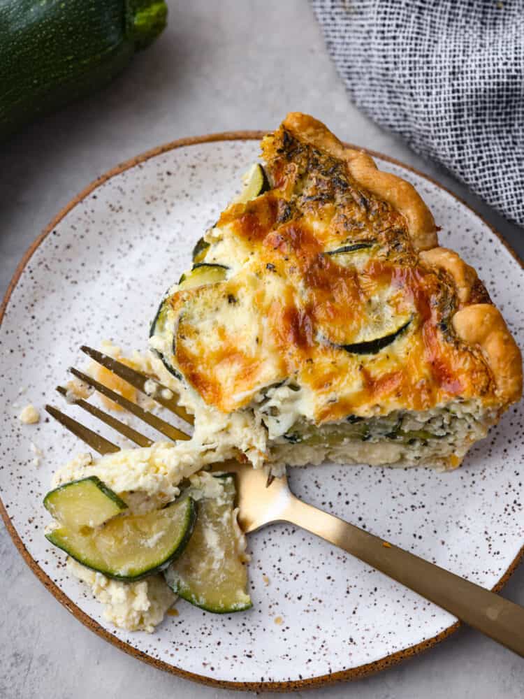 A slice of zucchini quiche on a plate with a fork cutting out a piece to eat.