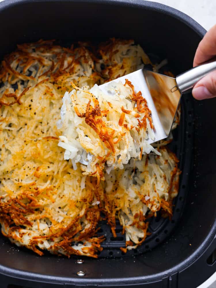 Close up view of cooked air fryer hash browns in the air fryer.  A spatula is lifting up a portion of hash browns.