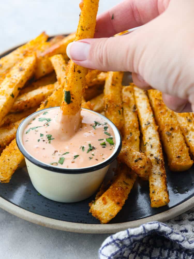 Close up view of a jicama fry being dipped into dipping sauce on a dark gray plate.  The dip is garnished with fresh parsley, and a blue and white checkered towel is next to the plate.