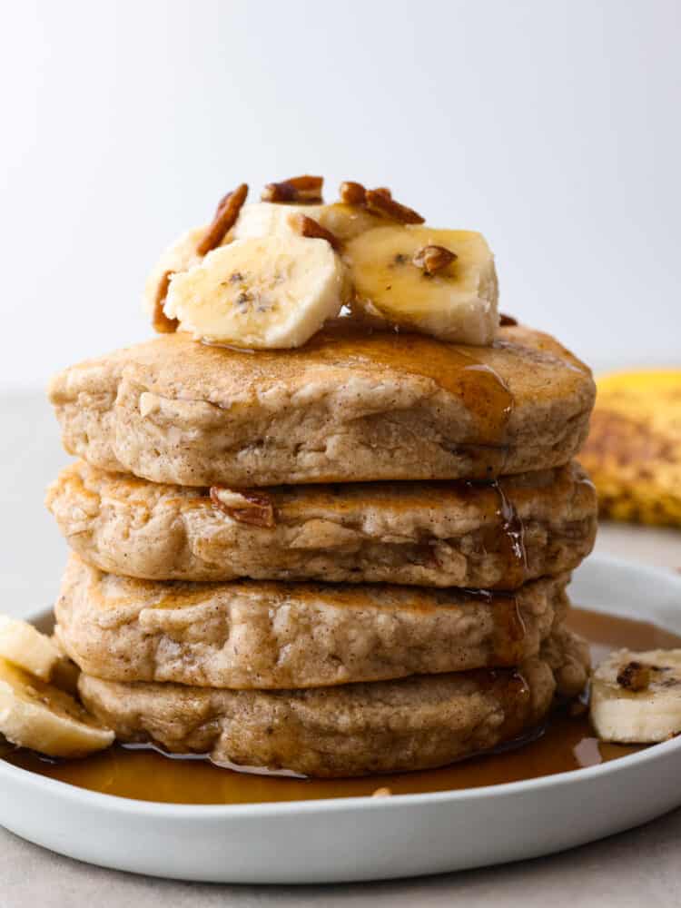 A stack of banana pancakes on a plate topped with fresh bananas and pecans. 