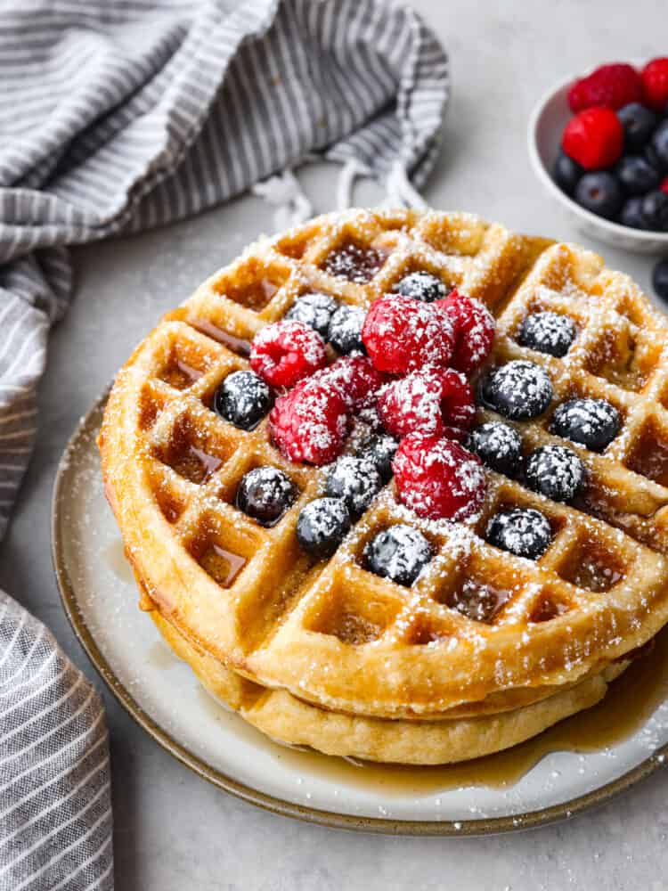 A stack of 2 waffles on a gray stoneware plate, garnished with berries and powdered sugar.