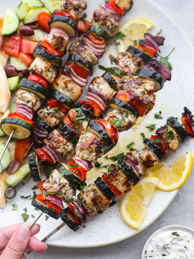 Close up photo of Greek chicken kabobs on a large white round platter.  Sliced pita bread, sliced cucumbers, diced tomatoes, and kalamata olives served on the side.  Sliced lemon garnished on the side with a small bowl of tzatziki sauce.  A hand is holding up one of the kabobs.