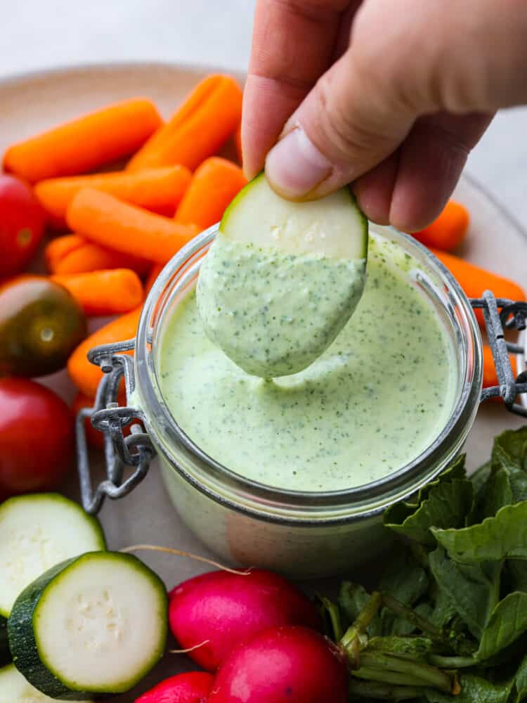 Top view photo of sliced zucchini being dipped into the jar of green goddess dressing.  Carrots, tomatoes, zucchini, and radishes are scattered around the jar of dressing.