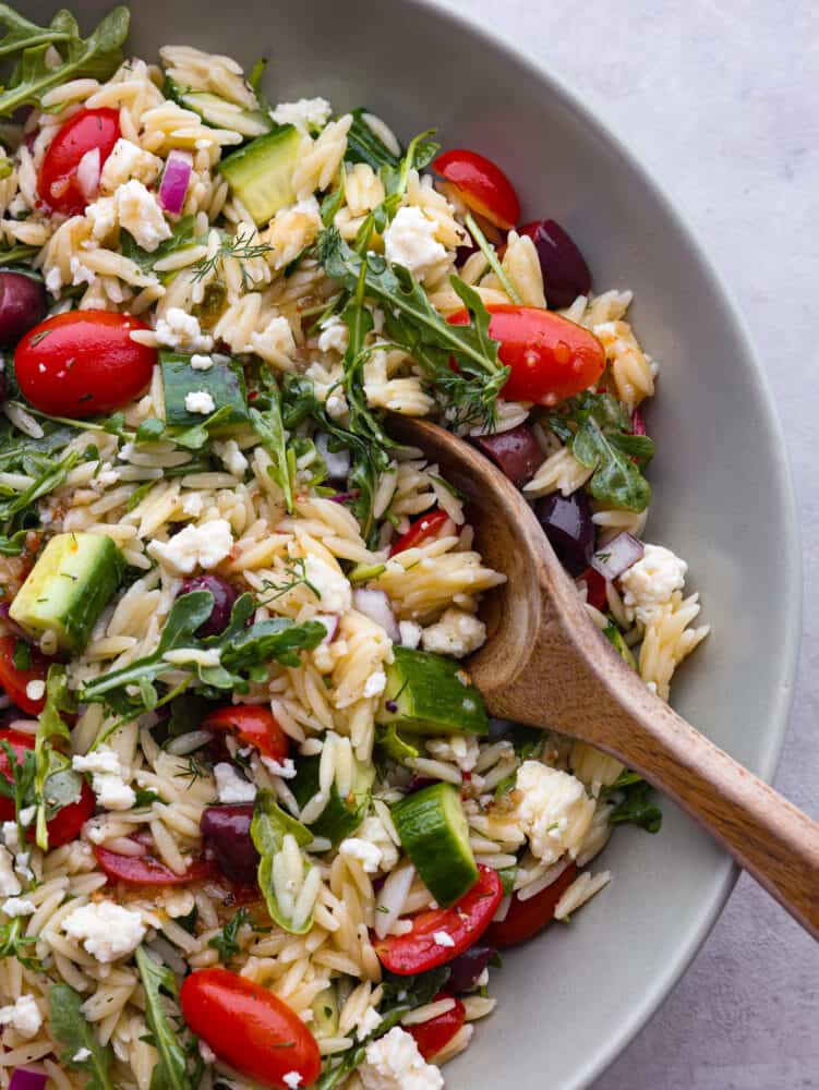 A close up of a bowl of Greek orzo salad with a wooden spoon. 