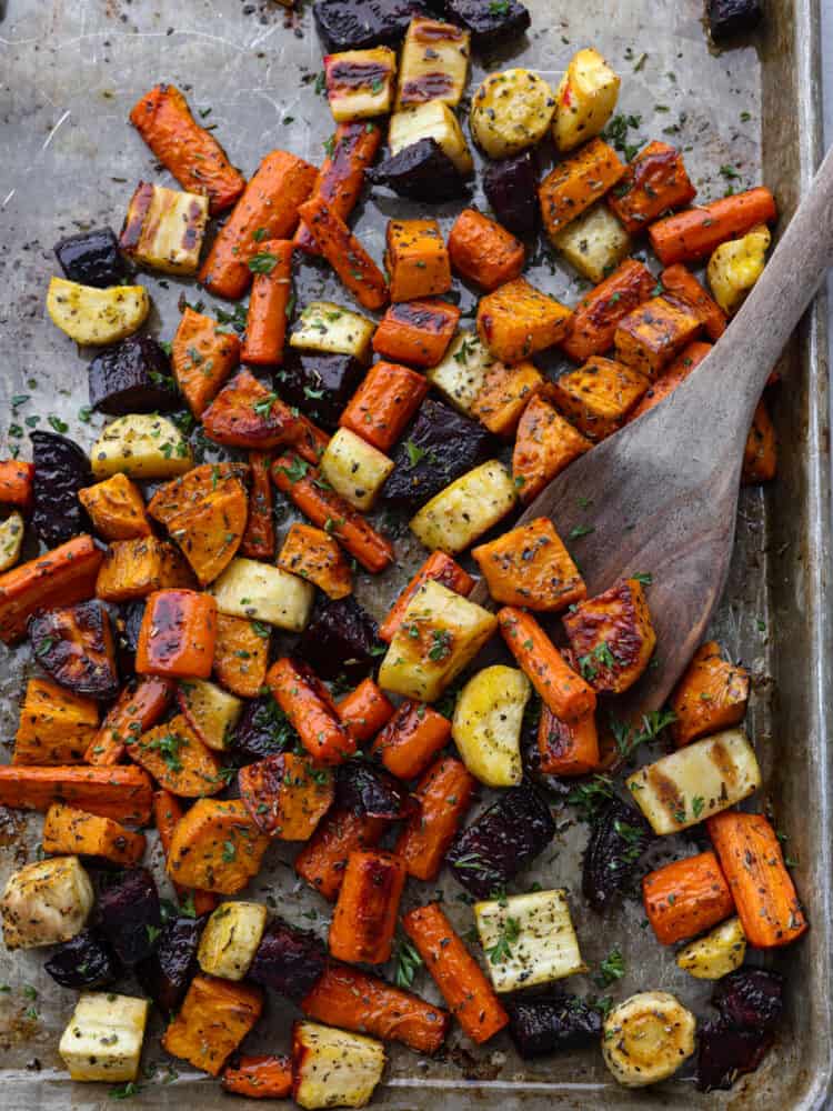 A baking sheet with roasted root vegetables being scooped up with a wooden spoon. 