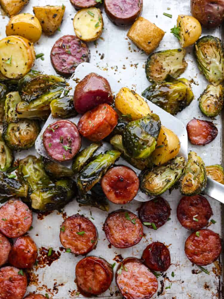 A close up of sausage and veggies on a sheet pan being scooped up with a spatula. 