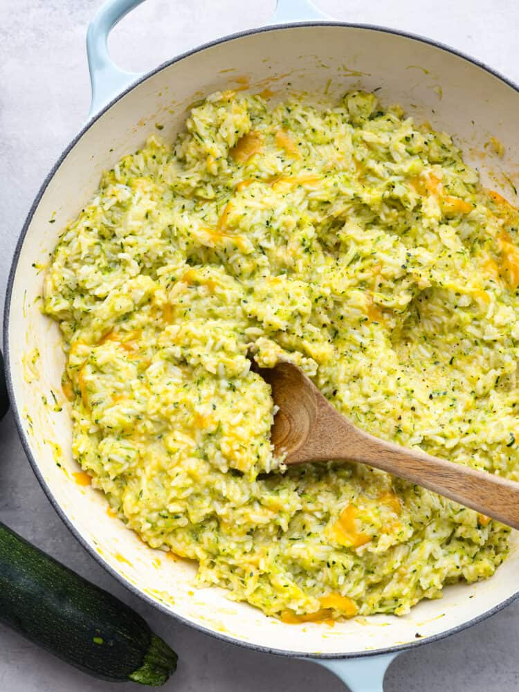 The top view of a pan filled with cooked cheesy zucchini rice and a wooden spoon. 