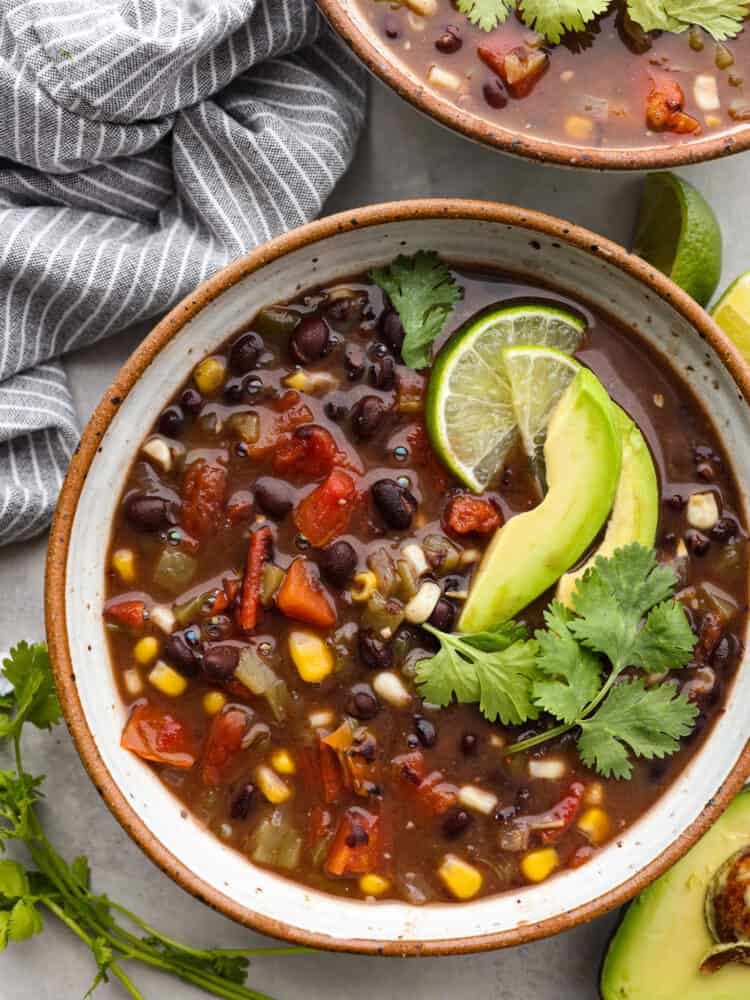 Top-down view of black bean soup in a speckled stoneware bowl, garnished with lime, avocado slices, and fresh cilantro.