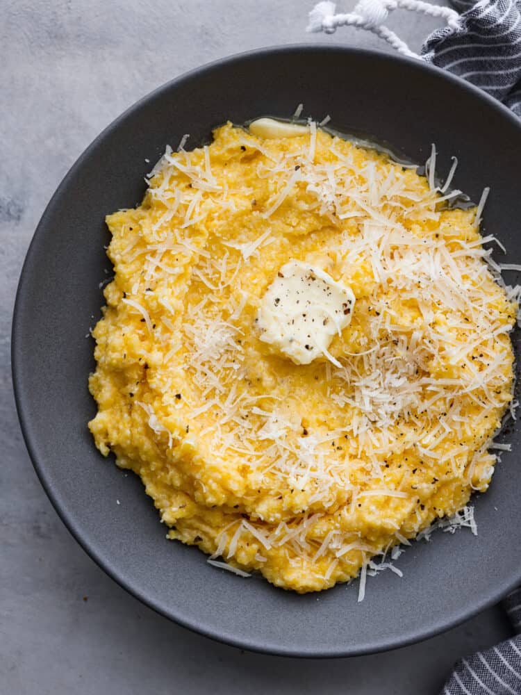 Creamy polenta in a gray bowl with butter, shredded parmesan cheese, and cracked black pepper garnished on top. A striped gray and white kitchen towel is next to the bowl of polenta.