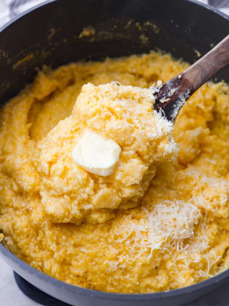 Close up view of creamy polenta in a large gray pot with butter and shredded parmesan cheese on top.  A wooden spoon is lifting up a spoonful of polenta. 