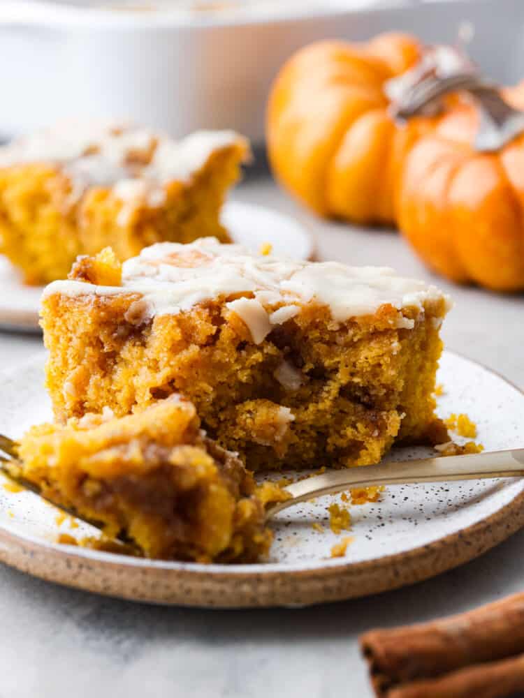 A slice of pumpkin cinnamon roll cake on a plate with a gold fork cutting a bite out of it. 