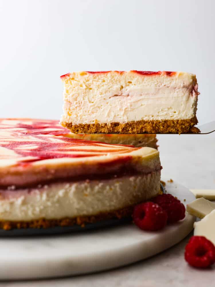 The side view of a full raspberry cheesecake with a slice being removed. 