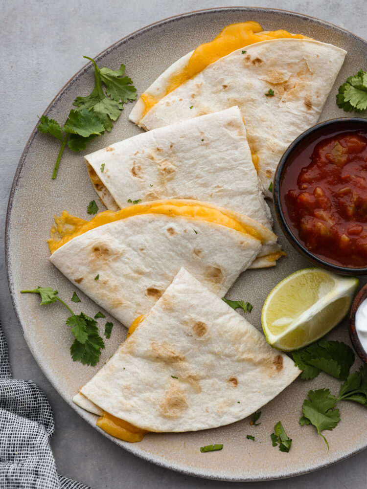 Top view of an air fryer quesadilla cut into triangles on a large gray plate.  A small bowl of salsa, cilantro leaves, and a lime wedge are also on the plate next to the quesadilla.