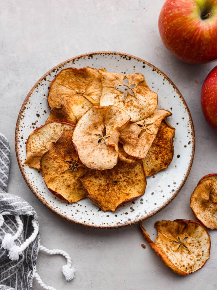 A pile of apple chips stacked on a small speckled plate.  Red apples and a towel are styled by the plate.