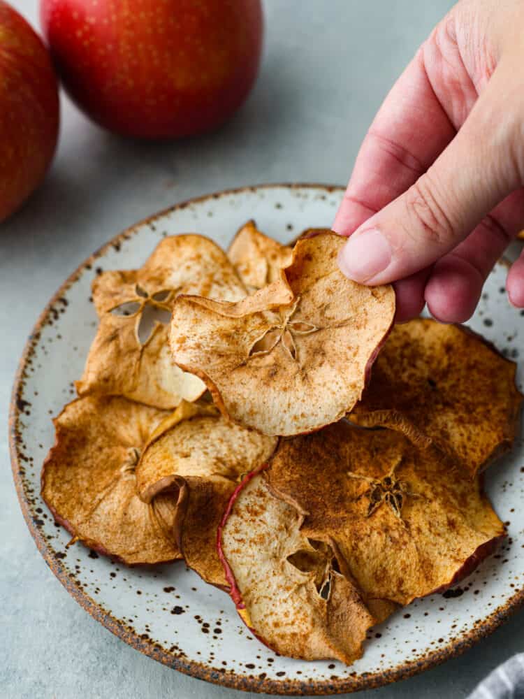 Close up photo of apple chips on a small plate.  A hand is picking up an apple chip.  Red apples are styled by the plate.