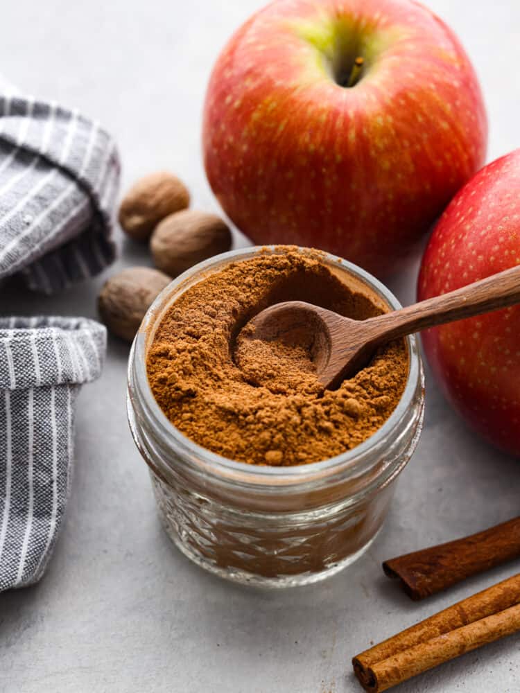 A clear jar of apple pie spice with a wooden spoon in it. There is a red apple in the background. 