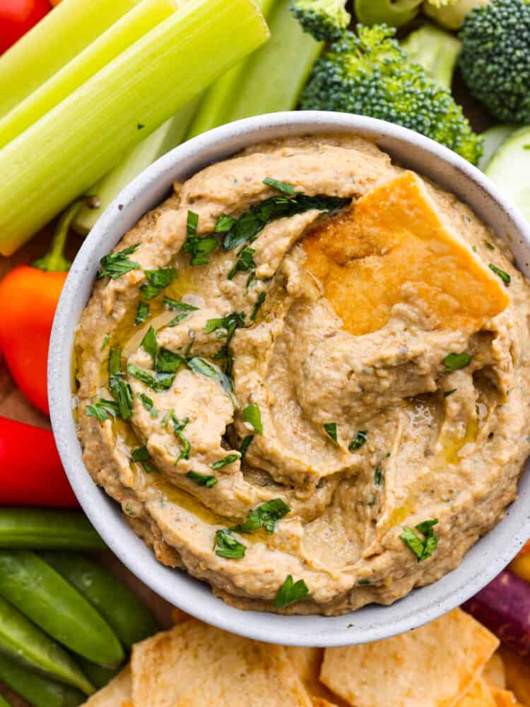 Closeup of baba ganoush in a white bowl, garnished with parsley and a pita chip.