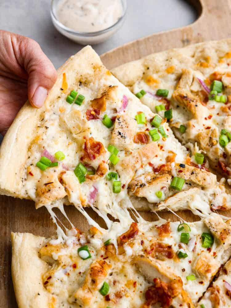 Closeup of a slice of chicken alfredo pizza garnished with bacon and chopped green onions being picked up.