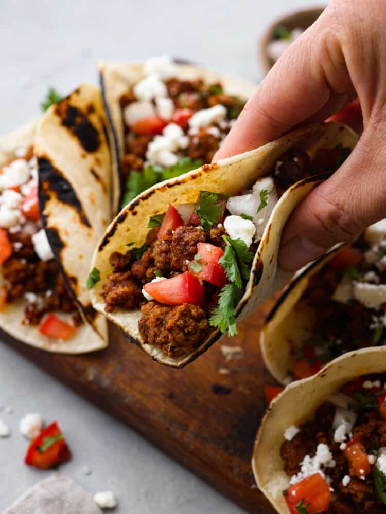 Chorizo meat inside of a flour tortilla with cilantro, tomato, onion and cheese being held by a hand above other tacos in the background.