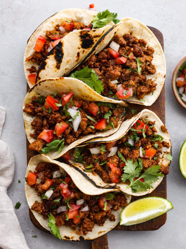 5 tacos served on a wooden board, garnished with fresh veggies, cilantro, and lime.