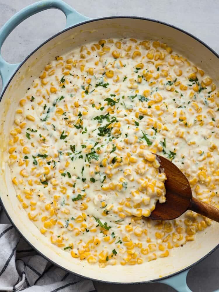 Top-down view of creamed corn, garnished with herbs in a white and blue skillet being stirred with a wooden spoon.